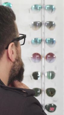 Olgun, sakallı bir adamın, bir optometri dükkanında canlı güneş gözlüğü dizisini incelerken sakalını nazikçe okşamasını yakalıyor. Düşünceli ifadesi, renkli gözlük görüntüsüyle birleşince, ilgi çekici bir sahne yaratıyor.