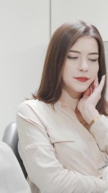 Endişeli bir ifadeyle genç bir kadın yanağına hafifçe dokunuyor, gösterişli, modern bir diş kliniğinde otururken diş ağrısını işaret ediyor. Bu canlı sahne, dişçi rahatsızlığının endişesini özetliyor, profesyonel bakımla karşılaştırıldığında...