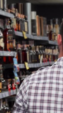 Dikiz aynasından çekilen bir görüntüde, derin bir şekilde süpermarket raflarındaki göz kamaştırıcı şarap şişelerinden birini seçmeye odaklanmış bir adam görülüyor. Şişelerin canlı renkleri parlak mağaza ışıklarının altında parıldıyor.