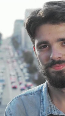 Neşeli, sakallı genç bir adam, renkli arabalar ve şehir hayatı ile dolu canlı bir şehir manzarası çerçeveli kameraya gülümserken mutluluk saçıyor. Bu şık hippi kendine güveni ve rahatlığı simgeliyor.