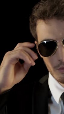 Özel dikim siyah takım elbiseli, çarpıcı, beyaz bir adam havacı güneş gözlüklerinin üzerinde kendine güven ve entrika saçıyor. Etkileyici bakışları, dramatik bir siyah arkaplan tarafından çerçevelenmiş, şık bir gizli ajanın özünü yakalıyor.