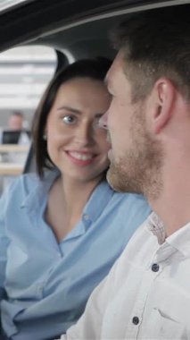 Genç bir çift, gülümseyen beyaz bir erkek ve kadın, galerideki şık bir arabanın içinde samimi bir konuşma yapıyorlar. Gözleri bir bağlantı kıvılcımında kesişiyor, sergi salonunun parlak, modern tasarımı ile çevrili, geleceklerini öngörüyorlar.