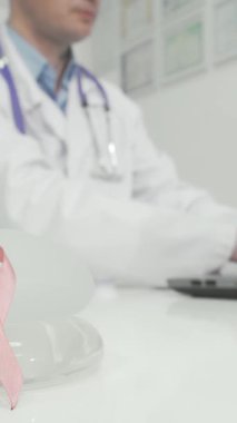Çarpıcı bir sahnede göğüs kanseri farkındalığının güçlü bir sembolü olan pembe kurdele parıldayan bir doktor masasında silikon göğüs implantlarının yanında yer alıyor. Huzurlu ortam, sağlığın, önlemenin ve