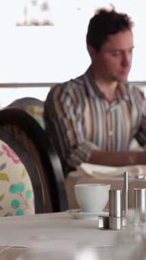 Şık bir kafede, sıradan giyinmiş genç bir iş adamı tabletiyle etkileşime girerek lezzetli bir yemeğin tadını çıkarıyor. Zarif masa dekorlarıyla süslenmiş şık bir ortam ve arka planda bir lokanta, mükemmel bir şekilde birleşiyor ve...