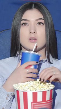 Dalgın Asyalı bir kadın canlı bir tiyatroda tek başına oturup patlamış mısır ve taze gazozun tadını çıkarırken titreyen ekrandan büyülenir. Canlı mavi koltuklar ve ekranın sıcak parıltısı davetkar bir ortam yaratıyor.