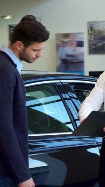 Gıcır gıcır beyaz gömlekli kendine güvenen sarışın bir satıcı her ikisi de sıkı bir tokalaşmayla meşgul şık esmer sakallı bir müşteriye panoda ayrıntıları sunuyor. Parlak siyah bir arabanın parlak arka planına göre bu dinamik sahne ışıldıyor.