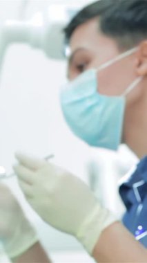 Maske ve eldiven takmış çalışkan genç bir dişçi, parlak ve steril bir klinikte hassas aletler kullanarak hastaların dişlerini titizlikle kontrol eder. Işıldayan diş aletleri ve odaklanmış ışıklandırma profesyonellik atmosferi yaratarak,