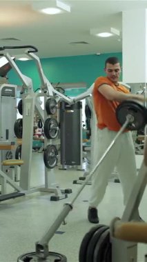 Orta Doğulu etnik kökenli kararlı bir genç, güçlü bir spor salonunda halter kaldırıyor ve göğsü ve pazıları üzerinde çalışırken gücünü ve dikkatini topluyor. Renkli duvarlar ve spor aletleriyle donatılmış modern spor salonu.