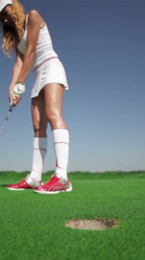 Odaklanmış genç beyaz bir kadın golf topunu delikten çıkarmak için zarifçe eğilir ve yemyeşil sahada yeteneğini sergiler. Şık beyaz bir kıyafet giymiş, canlı kızıl saçları parlak mavi gökyüzünün altında parlıyor,