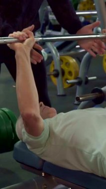 Odaklanmış Kafkasyalı üst düzey bir adam dinamik bir spor salonunda destekleyici genç bir erkek antrenörün rehberliğinde tutkulu bir şekilde bir halteri kaldırır. Canlı atmosfer renkli ağırlıklar ve modern ekipmanlarla canlıdır. Güçlendirici ruhu sergilerler.