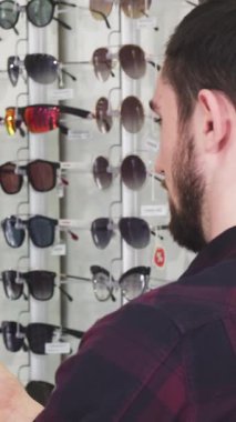 Genç bir erkek optometrist, gösterişli, iyi ışıklandırılmış bir gözlük dükkanında iki şık gözlük arasında müzakere ederken olgun sakallı bir adama hevesle yardım ediyor. Arka plan göz kamaştırıcı bir güneş gözlüğü ve optik çerçeveler dizisidir.