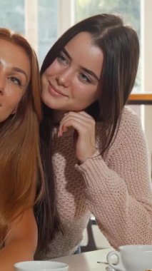 Canlı bir kahve dükkanında, birinin dalgalı kızıl saçlı, diğerinin parlak siyah bukleli iki neşeli genç kadın şakacı bir şekilde selfie çekiyorlar. Zarif hamur işleriyle bezenmiş ve yumuşak, sıcak ışıkla yıkanmış davetkar atmosfer mükemmel.