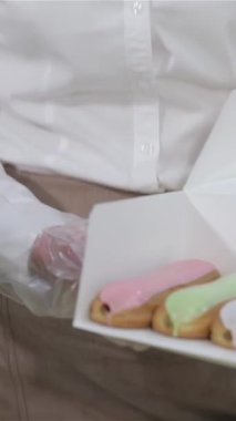 Canlı bir pastanede renkli, taze pişmiş keklerle dolu bir kutuyu sunarken kıtır kıtır beyaz gömlekli neşeli bir kadın. Sıcak gülüşü ve jöleli ikramların canlı renkleri davetkar bir atmosfer yaratıyor.