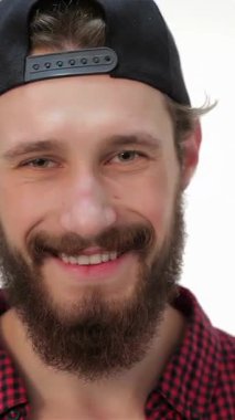 Moda bir şapka ve kareli gömlek giyen şık, genç sakallı bir adam, kameraya güvenli bir şekilde poz veriyor. Sıcak gülüşü ve hippi havası modern zarafet ve kişilik sergileyen davetkar bir atmosfer yaratıyor.