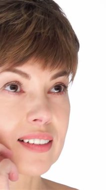 Orta yaşlı zarif bir kadın, kısa, şık saçlı, yanağına hafifçe dokunuyor, kendine güveni ve doğal güzelliği el değmemiş beyaz bir zemine yayıyor. Bu büyüleyici yakın çekim onun kusursuz cildini ve huzurlu ifadesini yakalıyor, izleyicileri davet ediyor.