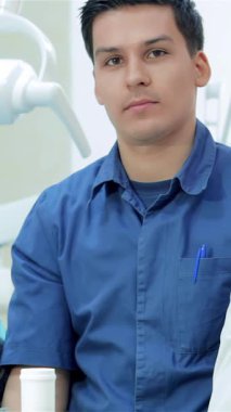 Parlak ve misafirperver bir diş hekiminde, kendine güvenen bir erkek dişçi üretken bir gün için hazır duruyor, profesyonellik ve samimiyet yayıyor. Odaklanmış ifadesi ve açık mavi üniforması hasta bakımına olan bağlılığını yansıtırken,