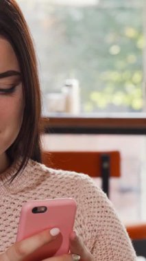 Sıcak, davetkar bir kahve dükkanında akıllı telefonuyla meşgul olan uzun, dalgalı saçları olan neşeli bir genç kadın. Yeni pişmiş hamur tatlısı aroması ve rahat bir ortam ile çevrelenmiş, boş zamanlarının mükemmel bir karışımını oluşturuyor.