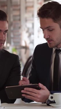 Şık bir kafede, iki iyi giyimli adam canlı bir restoranın davetkar atmosferiyle çevrili bir tableti incelerken yoğun bir şekilde stratejileri tartışıyorlar. Kararlı ifadeleri ve cilalı kıyafetleri profesyonelliği ve sinerjiyi simgeliyor.