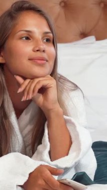 Uzun, dalgalı saçları olan ışıl ışıl beyaz bir bornoz giymiş lüks bir otel yatağında dinlenen sıcak gülümsemesi neşe ve rahatlık saçan genç bir kadın. Yumuşak bir yatak ve şık bir yatak başlığıyla güçlendirilmiş davetkar ortam mükemmel bir yatak başlığı yaratıyor.