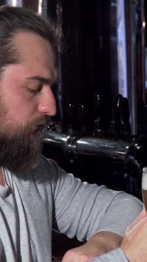 Düşünceli, gri süveterli sakallı bir adam barda oturmuş, düşünceli bir şekilde uzaktan bakarken köpüklü bira şişesinin tadını çıkarıyor. Yüzündeki ifade, yalnızlığın ve içselliğin özünü somutlaştıran dokunaklı bir karışımını ortaya çıkarıyor.