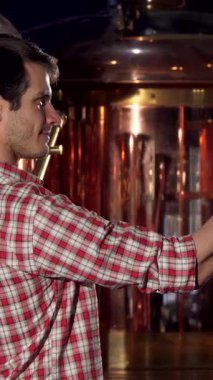 Şık, kareli bir tişört giymiş neşeli genç bir erkek bira imalatçısı, bir bardak altın birayı havada tutarken mutluluk saçıyor, canlı bir bira fabrikasında kendine güveni tamdır. Arkasındaki parıldayan bakır mayalama ekipmanı sıcaklığı arttırıyor.