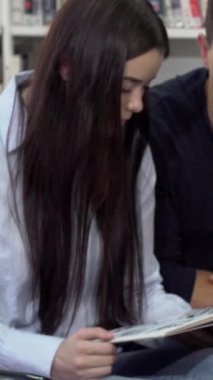 Neşeli bir kız öğrenci, canlı bir kütüphanede iş birliği yapan sınıf arkadaşları tarafından çevrelenmiş çalışma materyalleriyle uğraşırken neşe içinde ışıldıyor. Renkli kitap rafları ve hafif ışıklandırmalarla dolu sıcak, davetkar atmosfer,