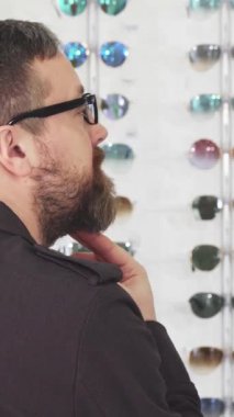 Sakallı, olgun bir adam, gözlükçülerin dükkanındaki göz kamaştırıcı güneş gözlüklerinden büyülenmiş bir şekilde omzunun üzerinden ışıldıyor. Neşeli tavırları heyecan saçıyor. Canlı seçimleri keşfedip alışveriş deneyimini gerçekleştiriyor.