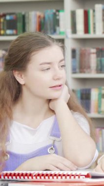 Üç genç kız, renkli kitaplıklarla dolu canlı bir okul kütüphanesinde canlı canlı sohbet ediyorlar. Gülüşleri ve paylaşılan gülümsemeleri, arkadaşlığın ve sevincin güzelliğini vurgulayan canlı bir atmosfer yaratıyor.