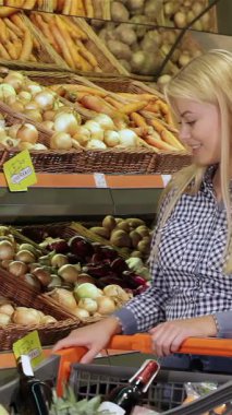 Modayı takip eden beyaz bir çift süpermarkette canlı bir alışveriş macerasının keyfini çıkarırken, şık sarışın kadın düşünceli partneri gözlemlerken taze, taze bir lahana almaya çalışıyor. Renkli sebze dizisi canlı bir zemin oluşturur.