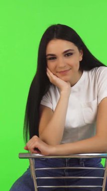 Uzun, dalgalı saçları olan ışıl ışıl genç bir kız modern bir sandalyede zarif bir şekilde oturuyor ve sıcak gülümsemesi sahneyi aydınlatıyor. Çarpıcı yeşil arka plan, onun neşeli ruhunu güçlendiriyor, enerjik ve davetkar bir atmosfer yaratıyor yaşam tarzı ve