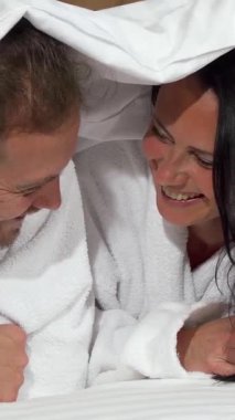 Pürüzsüz beyaz bir battaniyeye sarılmış neşeli olgun bir çift, yatakta yatarken bulaşıcı gülüşler ve sevgi dolu bakışlar paylaşıyor. Işıl ışıl gülüşleri ve neşeli ifadeleri samimi, samimi bir atmosfer yaratıyor. Aşkı ve bağı kutlamak için mükemmel.