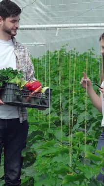 Bereketli yeşilliklerle dolu canlı bir serada genç bir adam renkli, taze toplanmış sebzelerle dolu bir sepeti heyecanla bir kız tarım uzmanına sunuyor. Gülüşleri sürdürülebilir çiftçiliğin ve işbirliğinin sevincini yayıyor.