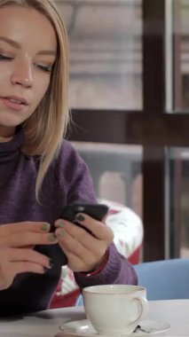 Tarz sahibi beyaz bir kız rahat bir kafede zarif bir şekilde oturur ve zarif parmaklarıyla akıllı telefonuyla rahatça dolaşır. Onun dalgalı sarı saçları ve rahat duruşu, dijital dünyaya kendini kaptırdıkça huzurlu bir hava yaratıyor.