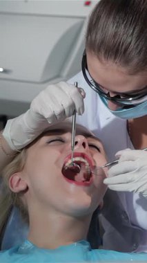 Gıcır gıcır beyaz önlük ve koruyucu giysi giyen kendini işine adamış genç bir dişçi kadın cerrah, hastaların dişlerini diş ve ayna kullanarak titizlikle inceliyor. Parlak, steril bir dişçi koltuğunda uzanan sarışın hastanın ağzı geniş.