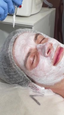 Huzurlu bir adam güzellik salonunda uzanır, yetenekli bir güzellik uzmanının titizlikle kremli bir yüz maskesi takması gibi gözleri mutlulukla kaplıdır. Bu rahatlatıcı tedavi, kendine bakmanın ve rahatlamanın özünü somutlaştırıyor.