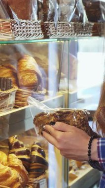 Uzun saçlı genç bir kadın, canlı bir fırında baştan çıkarıcı yeni pişirilmiş hamur işleri ile çevrili bir paket kurabiyeyi hevesle inceliyor. Bu canlı sahne, zevkli ikramları seçmenin heyecanını özetliyor, izleyicileri eğlenmeye davet ediyor.
