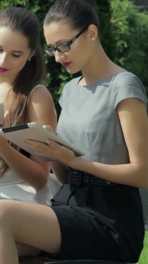 Canlı bir şehir parkında, iki şık iş kadını tabletlerine derinden odaklanırken, şık takım elbiseli üçüncü bir kadın da yakınlarda önemli bir telefon alıyor. Bu canlı sahne modern kurumsal hayatın nabzını somutlaştırıyor.