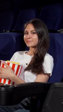 Çekici genç bir kadın rahat bir şekilde lüks bir sinema koltuğunda oturur ve elinde canlı çizgili patlamış mısır kovası tutarken yanındaki boş koltuğu işaret eder. Işıl ışıl gülüşü ve gevşek tavırları davetkar bir atmosfer yaratıyor.