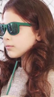 Dalgalı kestane rengi saçları olan hoş bir kız neşeli bir gözlük mağazasında neşe ve güven saçan şık bir güneş gözlüğü deniyor. Renkli çerçevelerin kaleydoskopu ile çevrelenmiş, çocukluk modasının özünü yakalıyor ve...