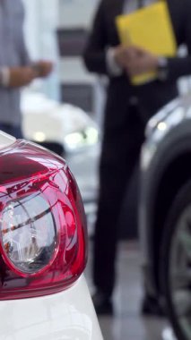 Bu büyüleyici yakın çekim modern bir arabanın parlak arkasını gözler önüne sererken, keskin giyimli bir satıcı da hareketli bir galeride potansiyel bir alıcıyı hevesle meşgul ediyor. Sahne, heyecanı ve profesyonelliği yansıtıyor.