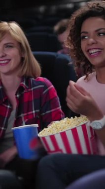 İki neşeli kadın, biri canlı kıvırcık saçlı ve diğeri şık kısa saçlı, rahat bir sinemada canlandırıcı bir filme dalmışken çizgili bir kova patlamış mısır paylaşıyorlar. Işıl ışıl gülümsemeleri ve rahat tavırları, izleyicileri davet eden sıcaklığı yayıyor.