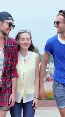 Üç şık genç arkadaş canlı bir şehir caddesinde el ele yürüyorlar, yüzleri neşe ve kahkaha ile parlıyor. Bu canlı sahne yaz ruhunu özetliyor. Renkli çiçeklere karşı moda kıyafetlerini sergiliyorlar.