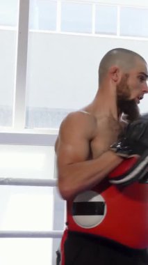 Azimli üstsüz bir erkek kickboksör, canlı bir spor salonunda kırmızı pedleri ustalıkla tutan odaklanmış koçuyla antrenman yaparken güçlü vuruşlar yapar. Her iki sporcu da sınırlarını zorlarken hava enerji ve hırsla dolu.