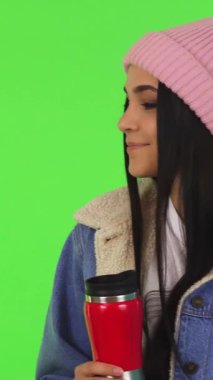 Şık bir kış ceketi ve yumuşak pembe bir bereyle giyinmiş hoş genç bir kadın, canlı yeşil renkli bir arka planda sıcak çayının tadını çıkarırken neşeyle ışıldıyor. Bu davetkar sahne sıcaklığı ve mutluluğu yansıtıyor, özünü mükemmel bir şekilde yakalıyor.