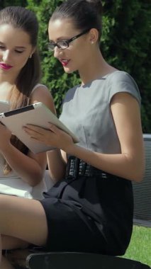 Canlı bir açık hava ortamında, iki şık iş kadını bir tablet üzerinde yoğun bir şekilde işbirliği yaparak profesyonellik ve güven yayarken, arka planda yer alan üçüncü bir kadın da canlı bir telefon görüşmesi yapıyor. Bu enerjik sahne,