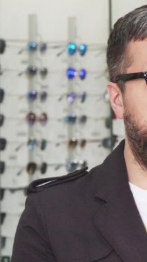 Sakallı bir adam şık bir optometriste dikilip, iki adet şık güneş gözlüğünü inceliyor. Renkli gözlüklerle çevrelenmiş, modern bir yetenek yayıyor, şık karar verme sanatını somutlaştırarak önceliklerini belirliyor.