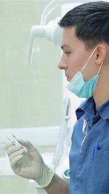 Kıtır mavi üniformalı ve maskeli gülümseyen bir dişçi, dişçi aletlerini dikkatlice seçer, profesyonellik ve samimiyet yayar. Parlak, steril klinik ortamı, genç bayan hasta için güven verici bir deneyim sağlayarak bakım atmosferini arttırır.