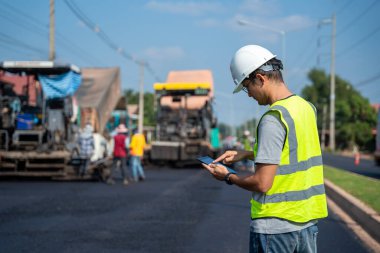 İnşaat mühendisi, inşaat mühendisi, asfalt kaldırım ve yol silindirli tablet ve atık kamyonu ve işçilerin yol geçmişi ile denetim ve tetkik altında güvenlik üniforması giyiyor.