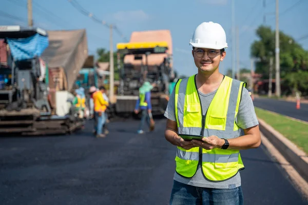 アスファルト舗装 道路ローラー ダンプトラックと労働者の建設道路の背景とタブレットで検査と調査の職場の下でアジアのハンサムな土木技術者 — ストック写真