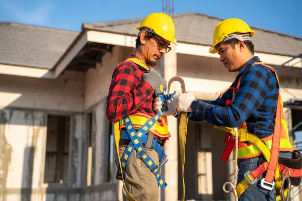 Asian two workers helping each other wearing fall protection suits with hooks at a construction site. safety hull structure working at height teamwork concept.